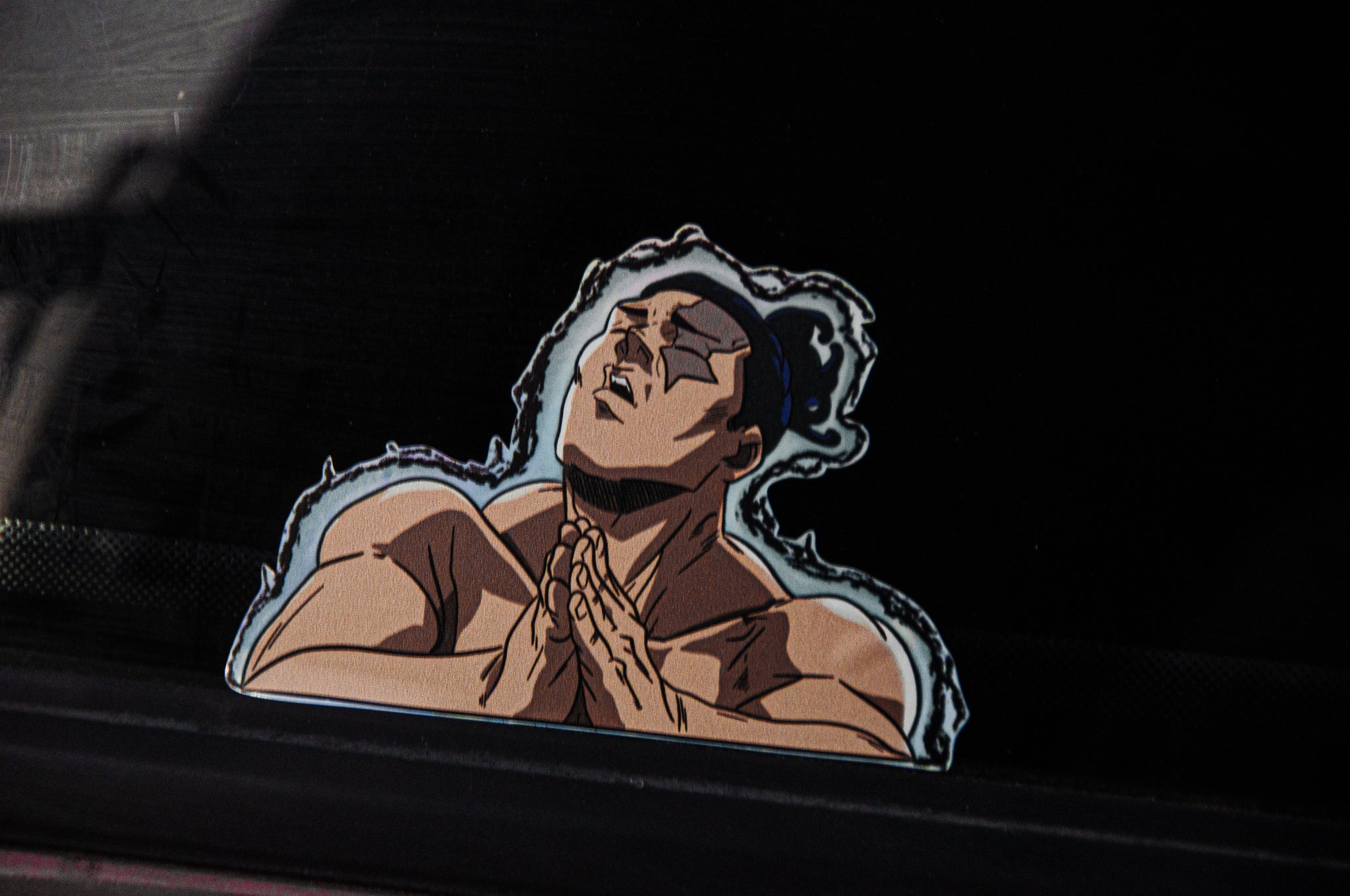 A sticker on a car window, the Aoi Todo Boogie!! Holographic Peeker Decal by Incompletegl, depicts an animated muscular figure reminiscent of Aoi Todo from Jujutsu Kaisen. The character has a serious expression, eyes closed, and hands clasped together in a prayer-like pose. The dark background makes the character stand out prominently.