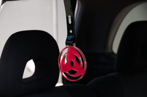 A red circular plastic Sharingan! | Tsurikawa from Incompletegl, resembling a JDM-inspired accessory, hangs from a black strap inside a car, positioned between the front seats. The background shows the interior roof and headrests of the vehicle's seats.