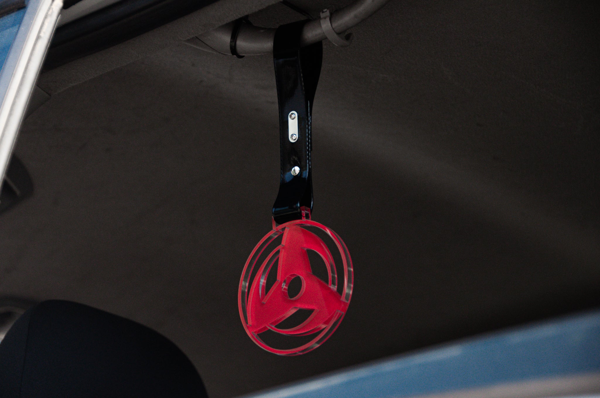 An Incompletegl Sharingan! | Tsurikawa, featuring a red and black ribbon with a round pendant resembling a wheel, hangs from the ceiling of a vehicle. The pendant, inspired by JDM designs and reminiscent of a Sharingan Tsurikawa, is attached to a black strap that is securely fastened to the overhead section of the car interior. This modern, abstract accessory adds an edgy touch to your ride.