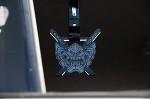 The Oni Mask! | Tsurikawa by Incompletegl is a rearview mirror accessory that showcases a dark-colored grinning Oni Mask with sharp teeth and menacing eyes, crossed by two swords in the background, adding a Japanese-inspired detail inside your vehicle.