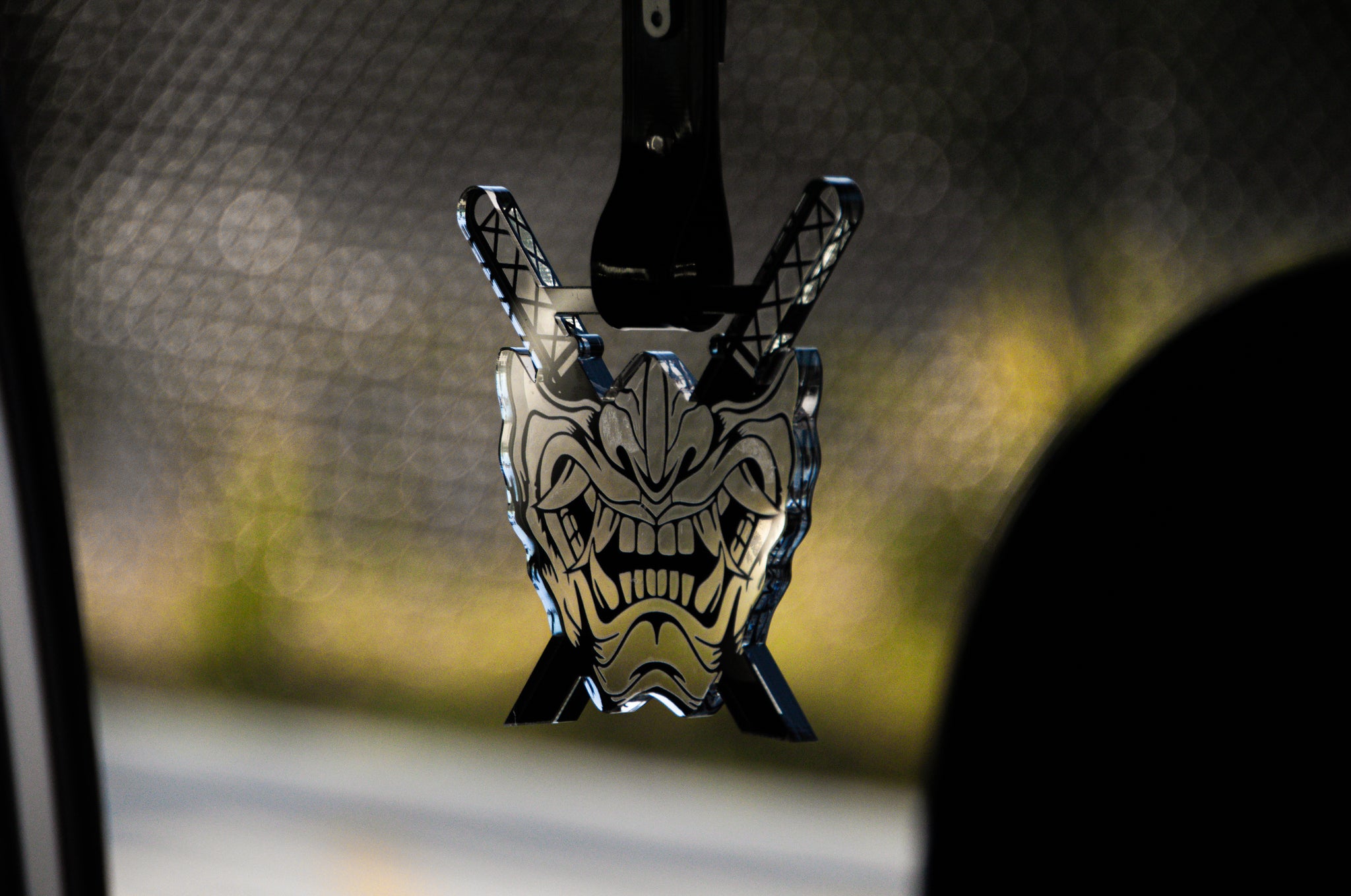 A close-up of Incompletegl's Oni Mask! | Tsurikawa rearview mirror hanging ornament inside a car, showcasing a meticulously detailed, menacing Oni Mask with an open mouth and sharp teeth. The background is out of focus, providing a faint glimpse of the road and greenery.