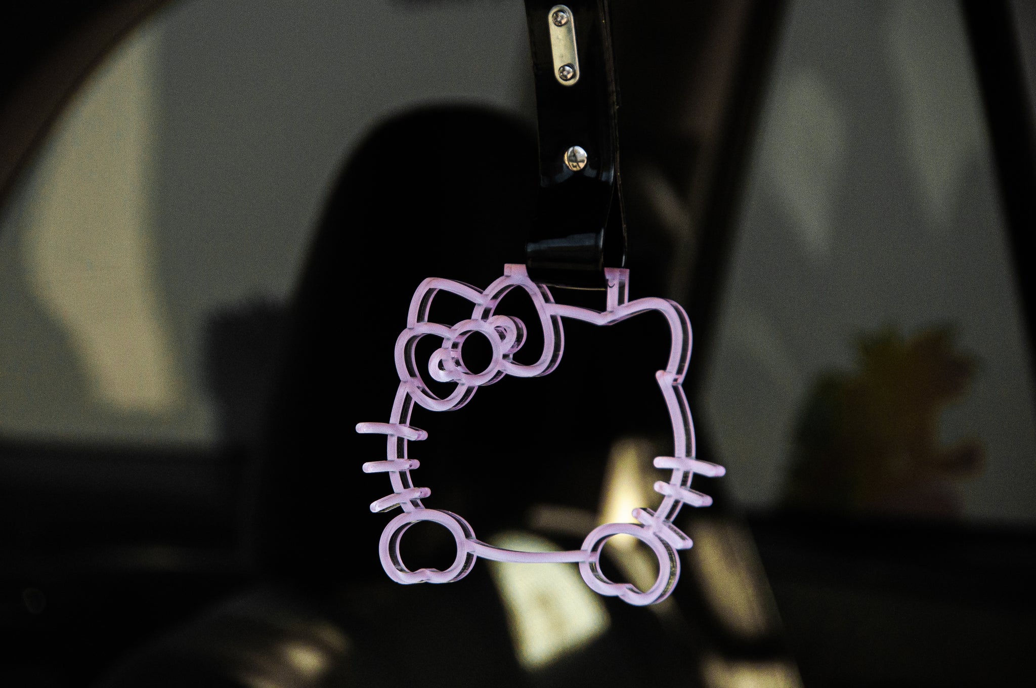 An adorable Hello Kitty! | Tsurikawa from the brand Incompletegl, shaped like Hello Kitty's head and crafted from a transparent purple material, dangles in a car as a rearview mirror charm. This JDM accessory showcases a cute bow on one ear, with the background displaying a blurred view of the car’s interior.