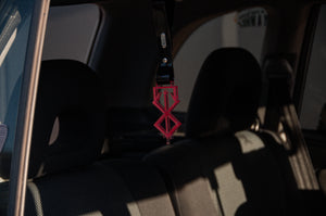 A pink geometric air freshener called "Brand of Sacrifice! | Tsurikawa" by Incompletegl hangs from the rearview mirror of a car, casting shadows on the seats. The interior appears dimly lit, with sunlight filtering through the windows, adding a subtle JDM touch.