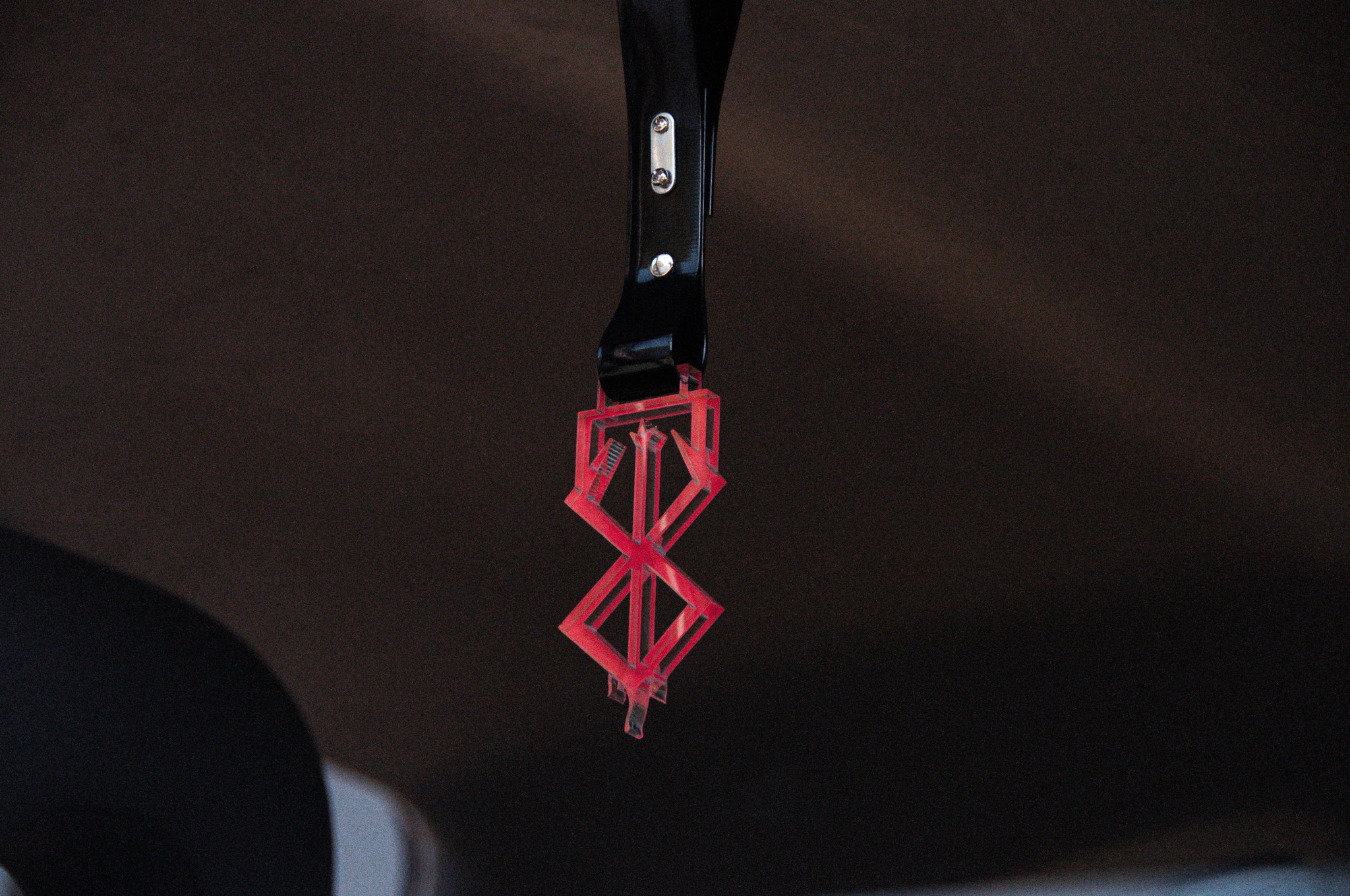 A red, geometric, double-diamond-shaped ornament with an acrylic finish, named the Brand of Sacrifice! | Tsurikawa from Incompletegl, is hanging from the black stem of a rearview mirror in a vehicle. The background is dark and blurred, highlighting the detailed design of this JDM touch hanging ornament.