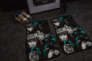 A Jujutsu Kaisen Mystery Pack from Incompletegl, which includes a pair of custom car floor mats adorned with anime-style characters featuring expressive designs and green accents, is placed on a dark carpet. Nearby, you can also find a pair of sneakers and an action figure from the anime series displayed against a framed item, evoking the essence of an exclusive bundle.