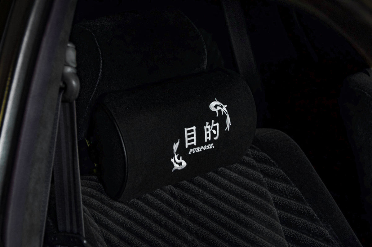 A car seat fitted with an Incompletegl Koi Fish Pillow headrest cushion, featuring a black cylindrical design adorned with white Japanese characters and the word "PURPOSE" embroidered in white underneath. The seat upholstery is black with a textured pattern.