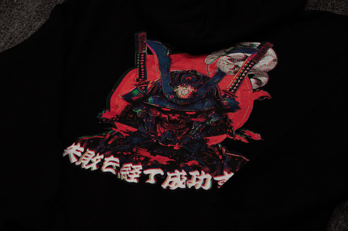 The Mecha Ronin | Oversized Hoodie by Incompletegl is a black hoodie featuring a robotic samurai helmet design in red, blue, and black tones. The background boasts a striking red circle with Japanese characters printed below the image. Perfectly capturing cyberpunk aesthetics, it’s laid out on a dark, textured surface.