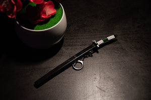 Close-up of an Incompletegl Demon Slayer Swords! Keychain featuring a black gravity-sheath design and a white handle, placed on a dark surface. A small round keyring is attached to the sheath. In the background, a white pot with vibrant red flowers and green artificial grass is partially visible, making it perfect for anime enthusiasts.