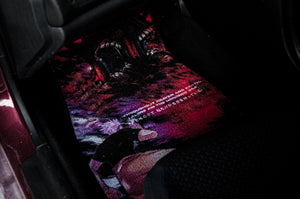 A close-up shot of a car's interior floor mat. The vibrant, detailed graphic on the Incompletegl "Gojo vs Sukuna, Domain Expansion!" Floor Mats depicts an intense red and purple scene reminiscent of Gojo vs Sukuna, with a menacing monster face and sharp teeth. Part of a black car seat and the vehicle's interior structure are also visible.