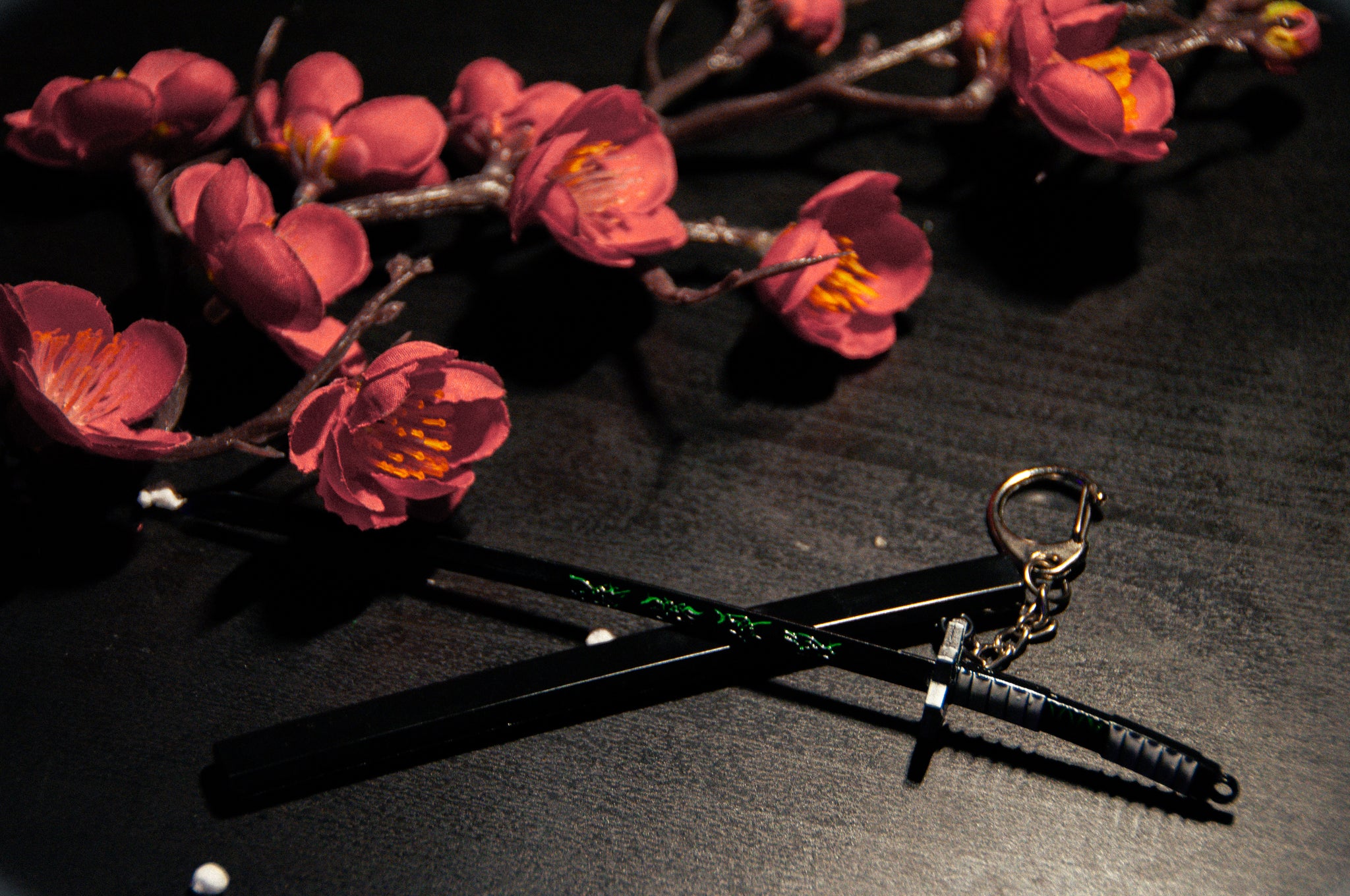 A pair of black chopsticks and a miniature Demon Slayer Swords! Keychain by Incompletegl are crossed over each other on a dark surface. Red cherry blossom branches with pink flowers are placed above them. The scene is lit subtly, creating a delicate and artistic composition perfect for anime enthusiasts.