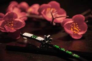 A miniature katana sword from the Incompletegl Demon Slayer Swords! Keychains collection, featuring a black handle with green accents, is placed on a black surface against the backdrop of pink cherry blossom flowers. The scene creates a striking contrast between delicate and powerful elements, making it a must-have for anime enthusiasts who appreciate intricate designs.