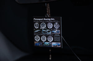 A close-up shot of a square Panasport Racing! air freshener by Incompletegl, showcasing various iconic wheels and photos of classic cars. The air freshener is suspended by a beaded string, dangling from the rearview mirror of a vehicle, set against a dark background.
