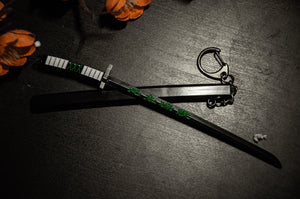 A detailed miniature black katana with green accents and a silver keychain attached lies on a dark surface. Orange decorative elements partially frame the top left corner of the image, making it perfect for anime enthusiasts seeking unique Demon Slayer Swords! | Keychains by Incompletegl.