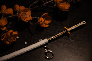 A decorative white katana with a detailed handle and an Incompletegl Demon Slayer Swords! keychain is placed on a dark surface. Beside it are orange flowers with a branch, adding a contrast of colors to the dark background. The scene has a moody and artistic ambiance, perfect for anime enthusiasts.