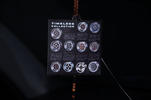 A close-up of a hanging decoration features a card labeled "Timeless Wheels! | Air Freshener" by Incompletegl. The card displays nine circular designs with descriptions in Japanese below each one. A string of brown beads is partially visible, connected to the card, echoing the precision found in JDM culture.