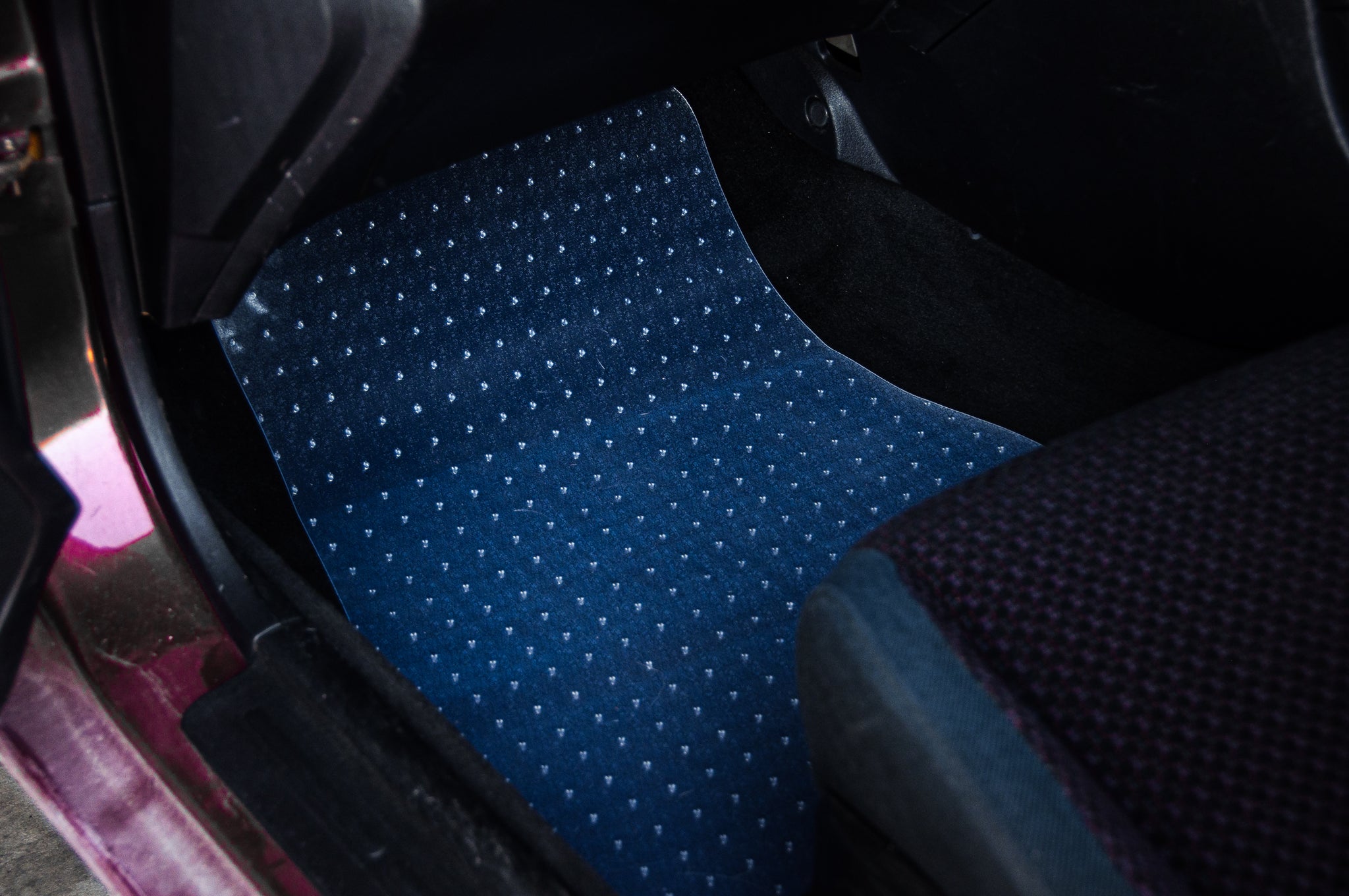 A close-up view of the front passenger floor area of a car reveals it is covered with *Floor Mat Protection!*'s blue, dotted PVC floor mat from *Incompletegl.*, perfectly designed for car interior protection. The edge of the car seat is partially visible to the right, highlighting the custom JDM mat's textured dotted pattern that stands out against the dark surroundings.