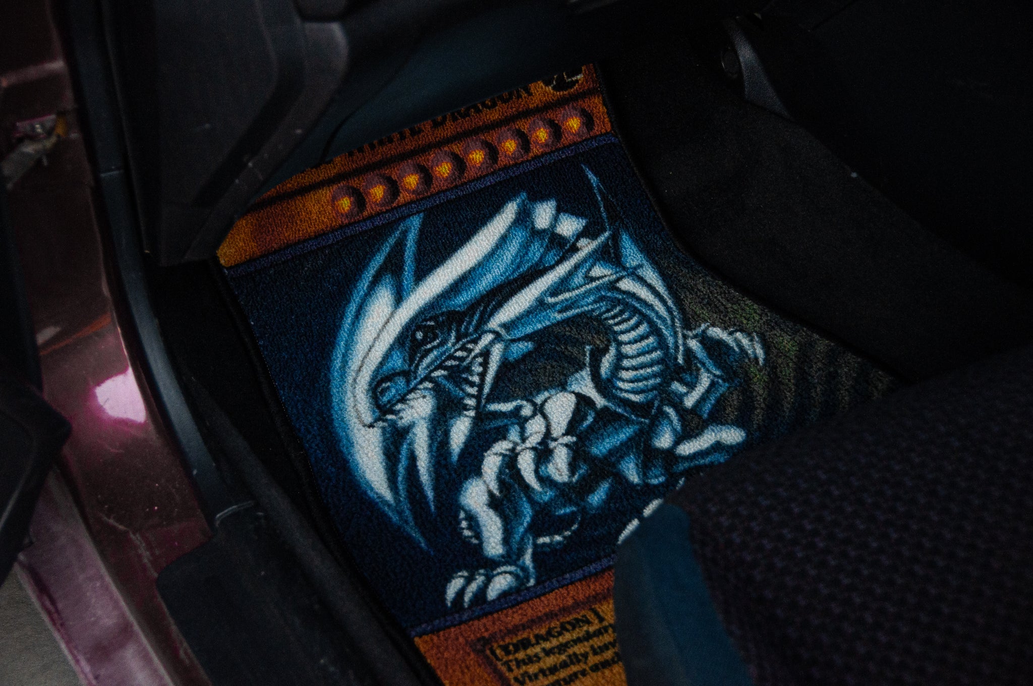 A car's footwell featuring the Blue Eyes!! | Floor Mats by Incompletegl, showcasing an anime-style blue dragon against an orange background. The dragon, reminiscent of Yu-Gi-Oh!'s Blue-Eyes White Dragon, displays striking details with sharp claws and a fierce expression. These floor mats also come with a non-slip backing for added safety.