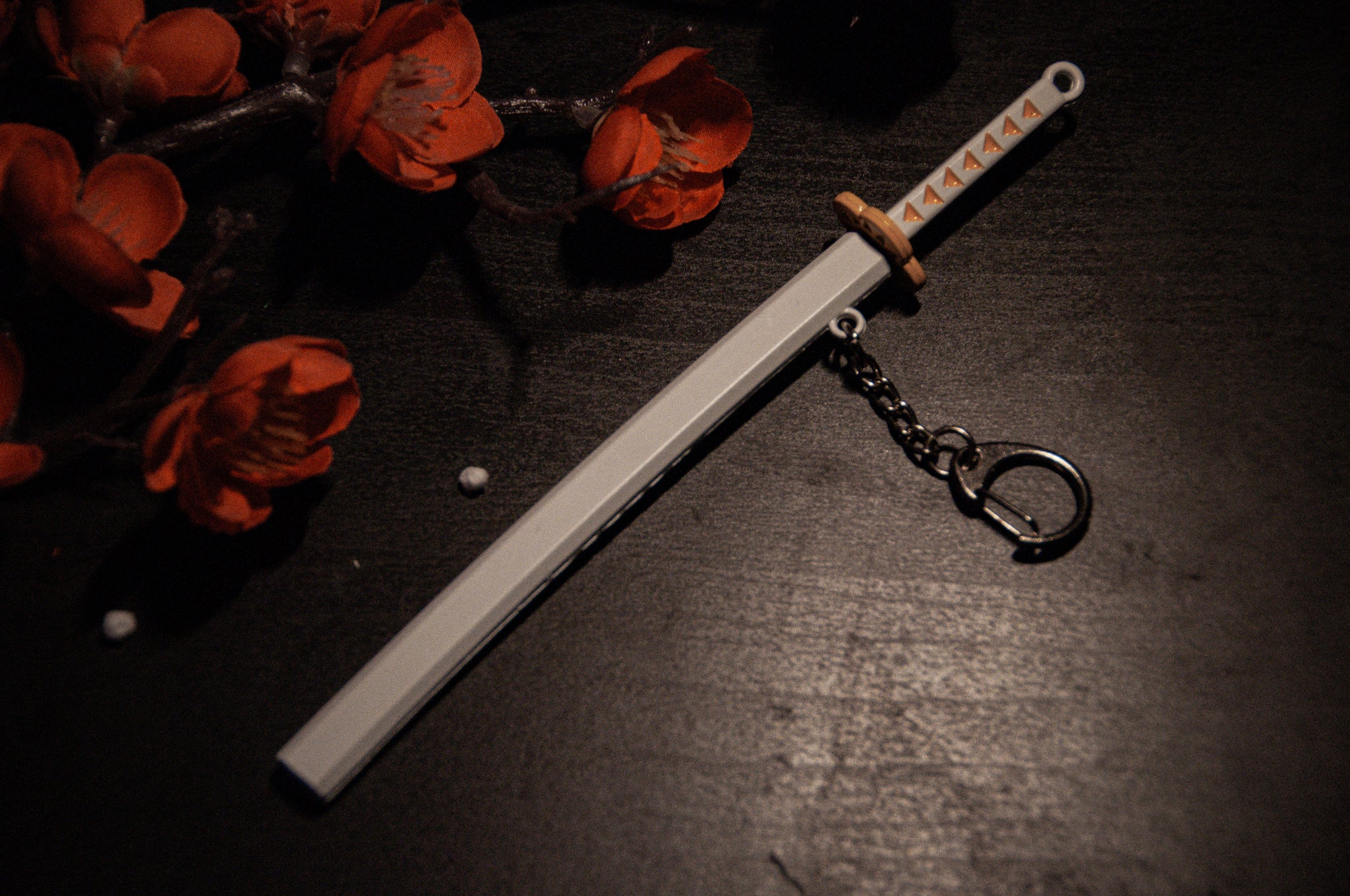 A miniature katana keychain with a white sheath and decorative handle lies on a dark wooden surface. Red artificial flowers occupy the upper left corner of the image. Perfect for anime enthusiasts, the Incompletegl Demon Slayer Swords! Keychain is attached to a small metal ring, ready to impress fans.