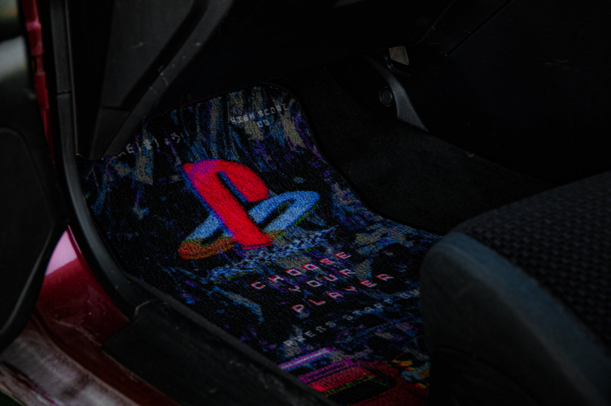 A close-up view of the "Retro Playstation! | Floor Mats" by Incompletegl, featuring colorful anime art and a retro gaming design. The mat prominently displays the classic PlayStation logo and the phrase "Choose Your Player" in vibrant colors, while the car's interior is partially visible around it.