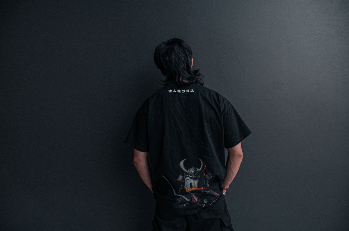 A person with long black hair is standing with their back to the camera, wearing an oversized DONALD DUCK x RONIN SUIT T-shirt by Incompletegl. The T-shirt features white text on the upper back and a graphic of Donald Duck in samurai attire at the bottom, set against a dark gray wall.