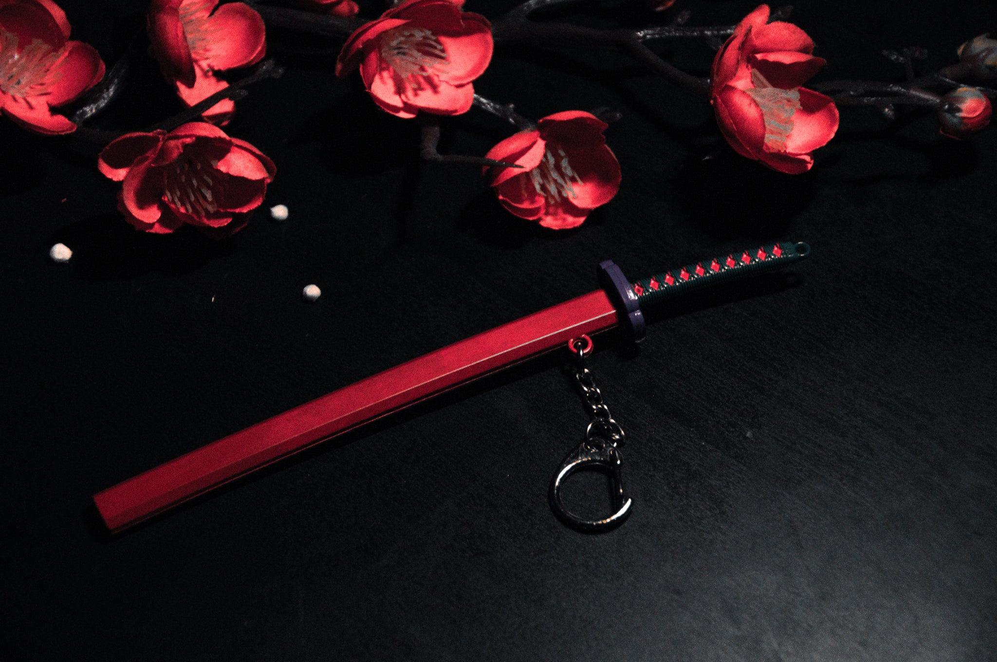 A small, decorative keychain from the Demon Slayer Swords! collection by Incompletegl lies on a dark surface. The keychain boasts a black and red handle, a small purple guard, and an elegant red scabbard with a gravity-sheath design. Surrounding the keychain are bright red flower blossoms—a must-have for anime enthusiasts!