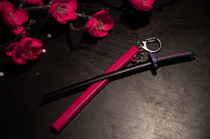 A Demon Slayer Swords! keychain from Incompletegl, featuring a miniature black blade and a vibrant handle, rests on a dark surface next to bright pink artificial cherry blossoms, captivating anime enthusiasts.