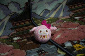 A plush toy featuring a pink bow is placed on a patterned blanket adorned with illustrations of pagodas, cherry blossoms, and cartoon characters. This charming accessory bears a resemblance to the Sanrio x Daruma Doll by Incompletegl, showcasing its round face, minimalistic features, and pink accents—making it an ideal addition for your car interior.