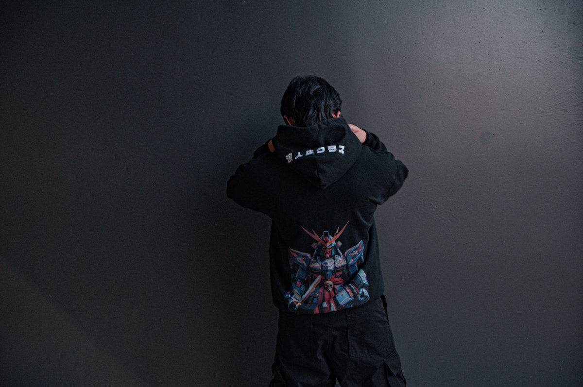 A person with short black hair is facing away from the camera, adjusting their GUNDAM x RONIN SUIT | Oversized Hoodie by Incompletegl. The back of the hoodie showcases vibrant artwork depicting a Gundam robot dressed in samurai attire, set against a plain dark grey wall.