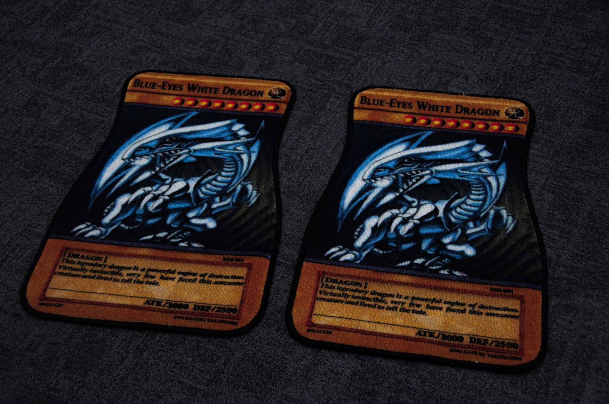 A photograph showcasing two Blue Eyes!! floor mats from the brand Incompletegl, placed on a dark gray surface with a non-slip backing. Both mats feature an illustration of a dragon with blue and white coloration, boasting identical stats and descriptions.