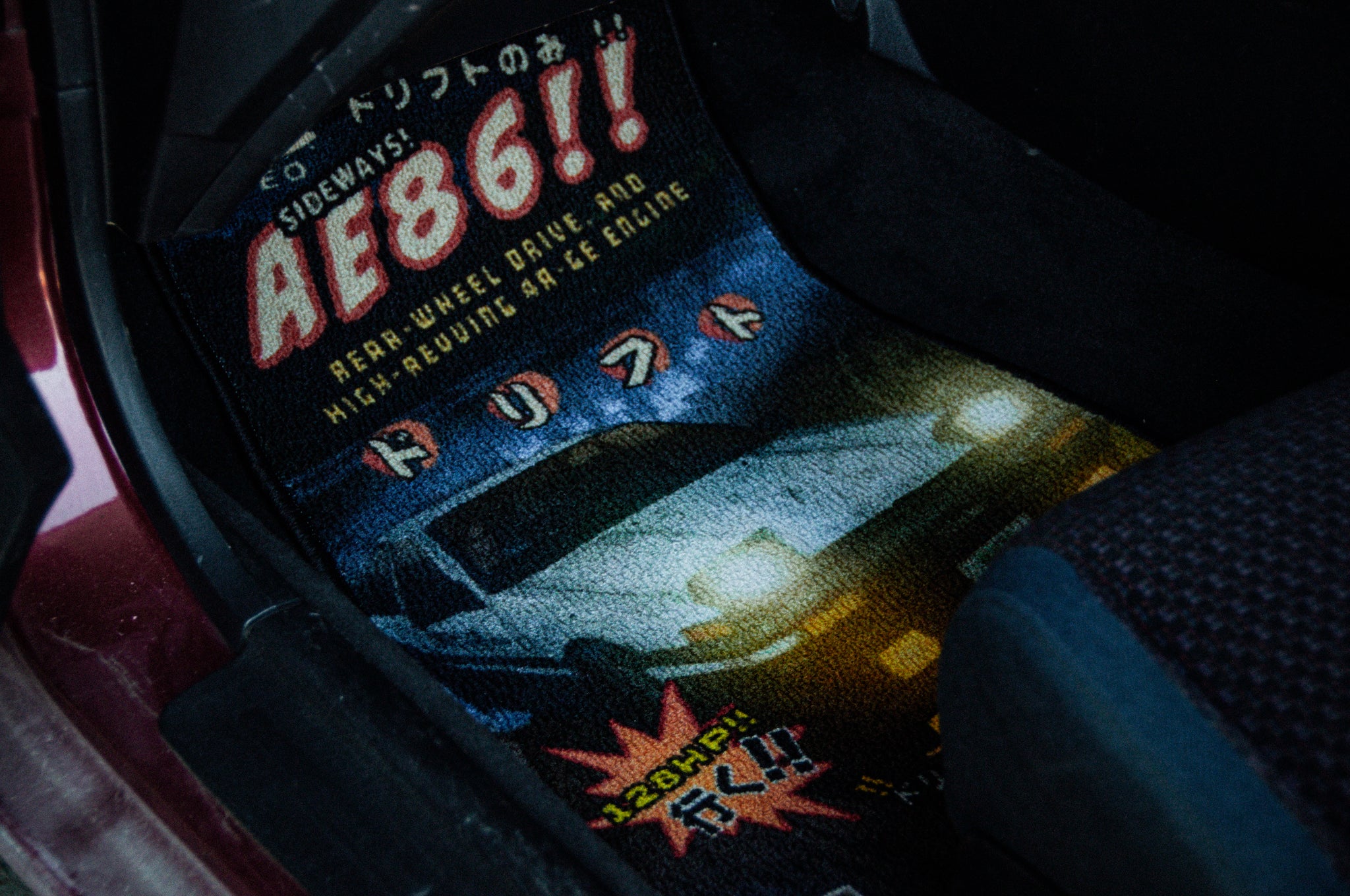 A close-up photo of an Initial D; Retro Style!! | Floor Mat inside a vehicle. The mat from Incompletegl showcases an illustration of the iconic AE86 with headlights on, accompanied by Japanese text and the bold red "AE86!" This design pays homage to JDM culture and celebrates the performance of the legendary AE86 Toyota model seen in Initial D.