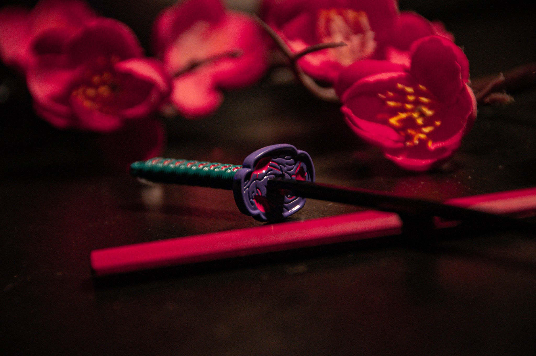 An exquisite small katana, identical to the "Demon Slayer Swords! | Keychains" by Incompletegl, with an ornate hilt and a green handle, rests on a vibrant red sheath against a black background. Surrounding the katana are radiant pink cherry blossoms, enhancing the traditional Japanese aesthetic that anime enthusiasts admire.