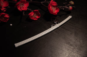 A Demon Slayer Swords! Keychain by Incompletegl, featuring a miniature samurai sword with a white blade and silver handle, rests on a dark surface. Above and to the left of the keychain are bright red artificial cherry blossoms on slender branches—an ideal setup for anime enthusiasts.