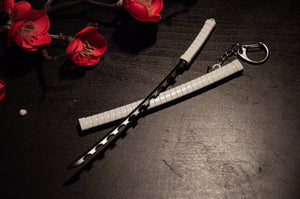 Close-up of two crossed miniature Demon Slayer Swords! | Keychains with white-wrapped handles, resting on a dark surface. These keychains, featuring a gravity-sheath design by Incompletegl, are accompanied by metal keychain loops. Bright red decorative flowers lie beside the swords—an essential addition for anime enthusiasts and collectors of Demon Slayer Sword Keychains.