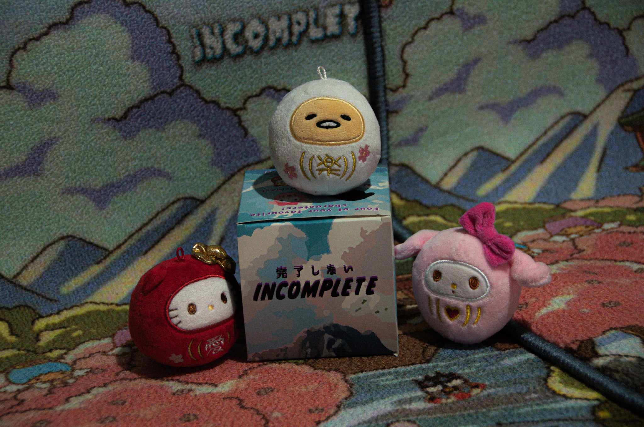 Three plush toys designed as round characters with simple facial features and robes are arranged on and around a box labeled "Incompletegl," reminiscent of the Sanrio x Daruma Doll Blind Box! The backdrop is colorful and abstract, featuring clouds and mountains, creating a whimsical scene similar to discovering treasures in a blind box.