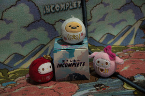 Three plush toys designed as round characters with simple facial features and robes are arranged on and around a box labeled "Incompletegl," reminiscent of the Sanrio x Daruma Doll Blind Box! The backdrop is colorful and abstract, featuring clouds and mountains, creating a whimsical scene similar to discovering treasures in a blind box.