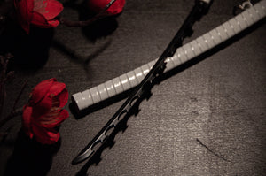 A close-up photo features the uniquely designed, serrated sword from the Demon Slayer Swords! | Keychains by Incompletegl, with a white-wrapped handle lying next to its matching gravity-sheath design scabbard on a dark surface. Bright red flowers are partially visible in the background, providing a contrasting splash of color for anime enthusiasts.