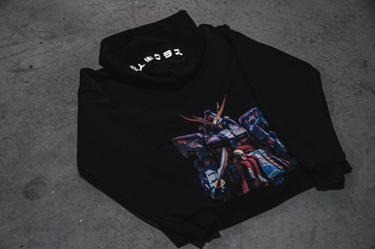 The GUNDAM x RONIN SUIT | Oversized Hoodie by Incompletegl features a black design with a folded cowl neck, adorned with a vibrant Gundam graphic on the front. Japanese text in white embellishes the cowl neck area. The hoodie is displayed on a gray concrete surface, merging modern style with samurai-inspired elements.