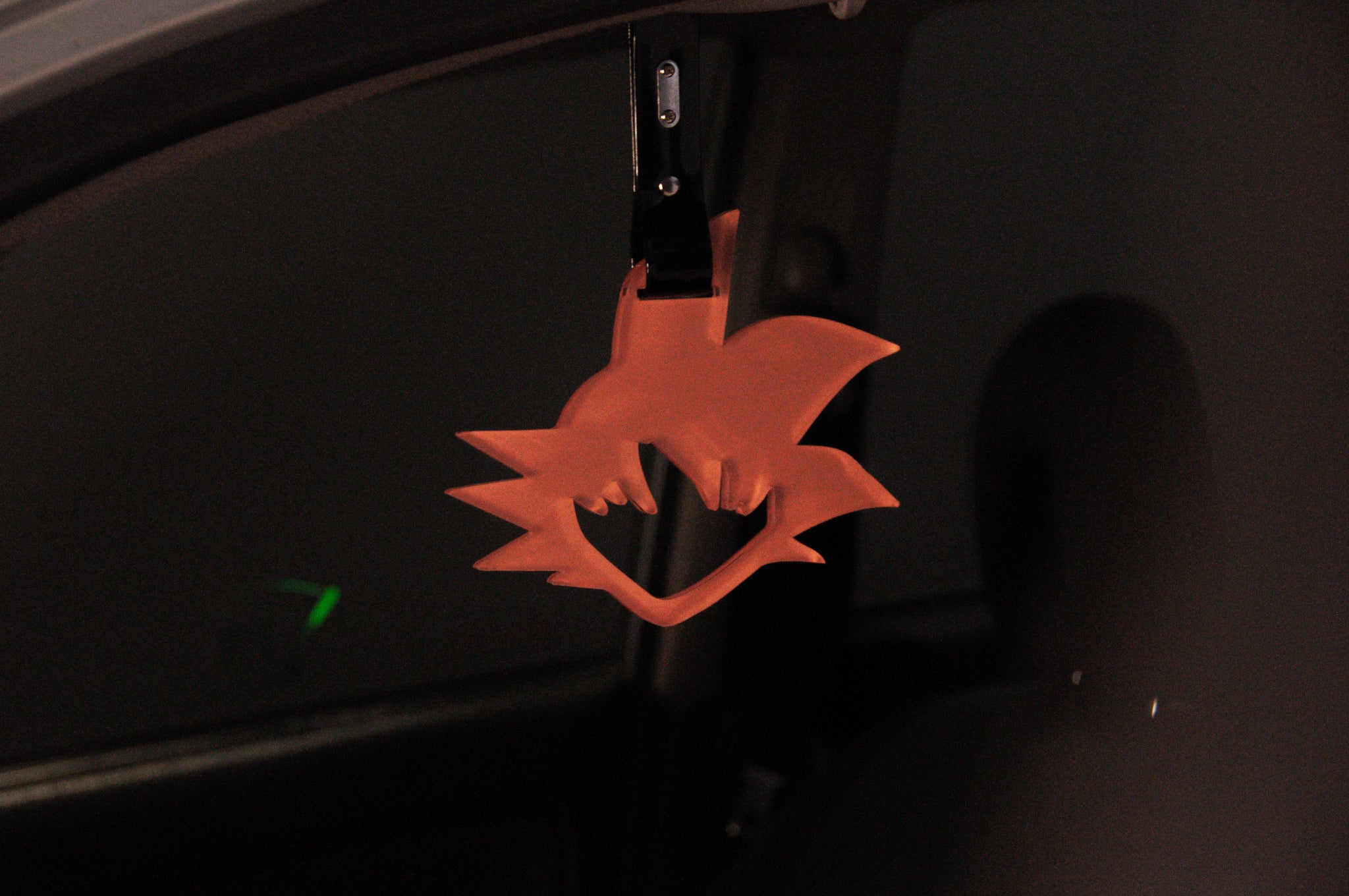 A dimly lit car interior showcases the striking detail of a red, stylized hanging ornament from the Goku! | Tsurikawa line by Incompletegl, resembling a fox or wolf head suspended from the rearview mirror. This Japanese car accessory prominently stands out against the dark background, echoing elements of JDM culture.