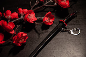 A close-up shot of a decorative black Demon Slayer sword keychain with a red and black hilt, resting on a dark surface under the brand name Incompletegl. Beside it lies a branch adorned with vibrant red artificial flowers. The scene features minimal lighting, drawing attention to the intricate katana keychain and flowers—a perfect display for anime enthusiasts who admire such artistry.