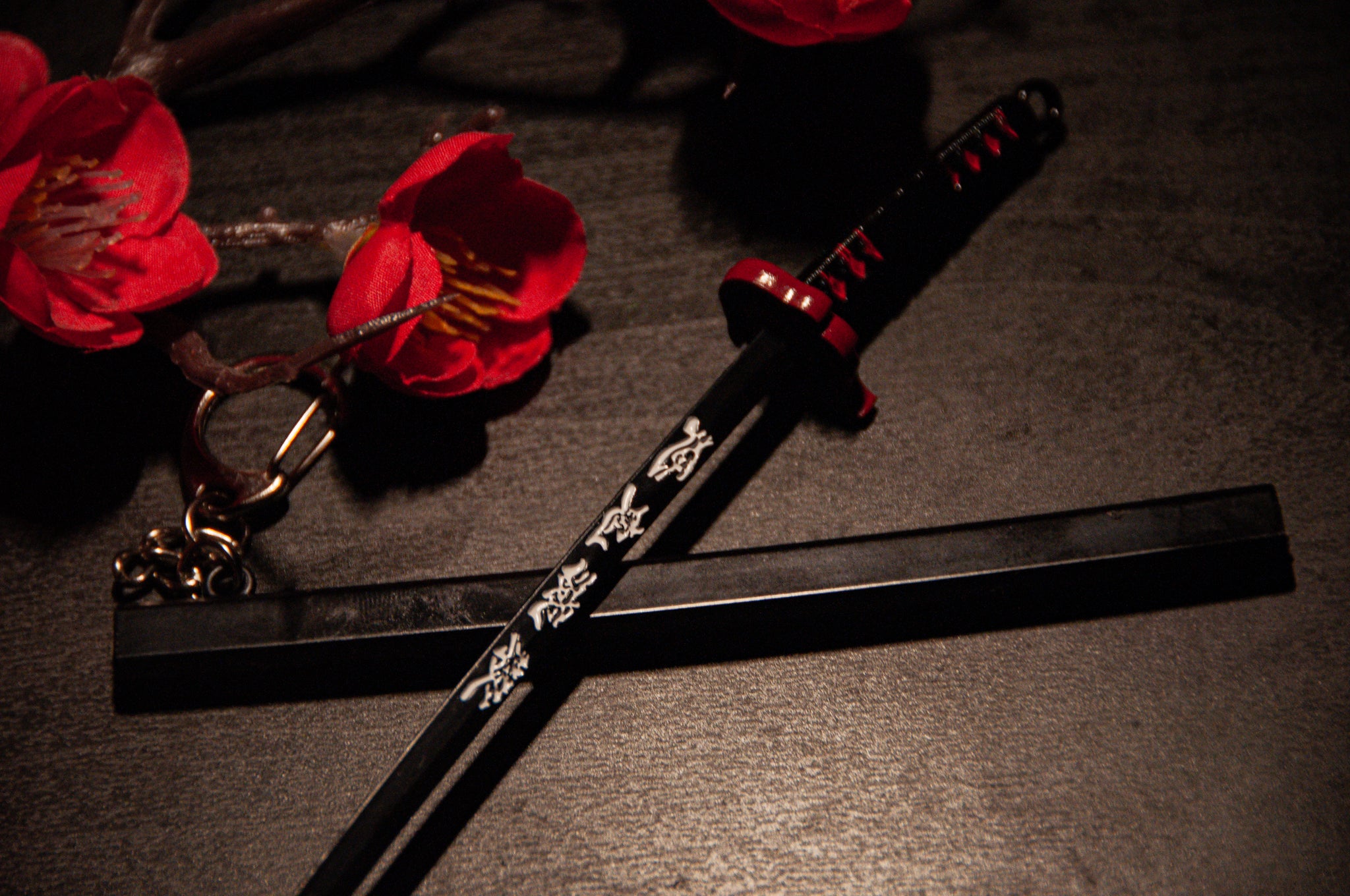 A black Demon Slayer sword keychain by Incompletegl, featuring intricate white designs on its blade and a gravity-sheath design, rests on a dark surface. The handle is adorned with red accents. Nearby, red flowers with yellow centers add a contrasting splash of color to the scene, making it perfect for anime enthusiasts.