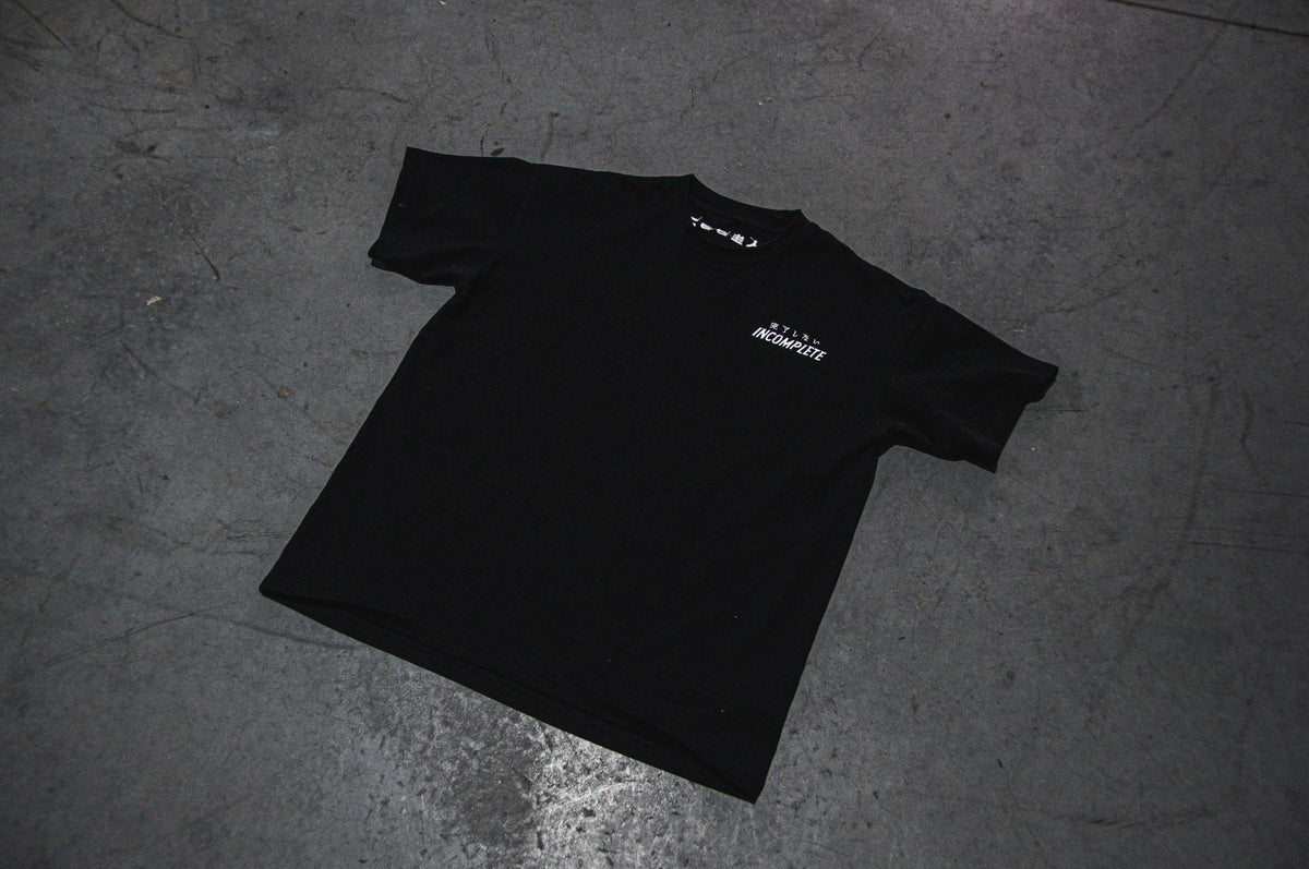 An Incompletegl DONALD DUCK x RONIN SUIT Oversized T-Shirt in black, featuring white text and small graphics on the upper left chest area, is laid flat on a gray concrete floor. The lighting casts a slight shadow around the T-shirt, accentuating its shape and texture—perfect for those embracing their inner Ronin Class warrior.
