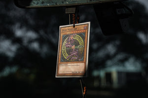 An air freshener featuring the Dark Magician from Yu-Gi-Oh!, branded as Incompletegl, is hanging from a rearview mirror in a car. The background is blurred, showing dark, out-of-focus trees and a hint of a building. The spellcaster-themed air freshener is suspended by a beaded string in an environment that appears dim and overcast.