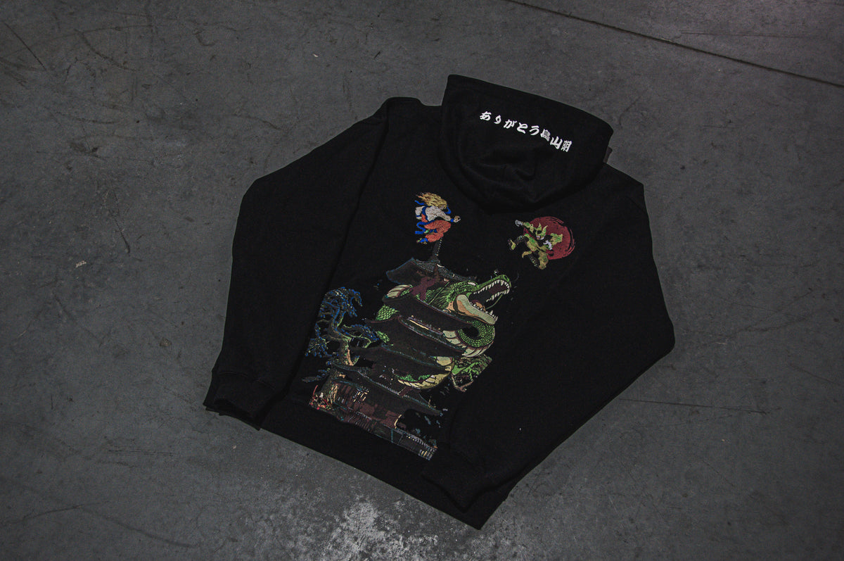 The Clash of Shenron x Ukiyo-e Style oversized hoodie from Incompletegl is a black garment showcasing a vibrant graphic design of a dragon and anime-like figures, paying tribute to Akira Toriyama. The back features Japanese characters on the collar, and the hoodie is displayed against a concrete floor.