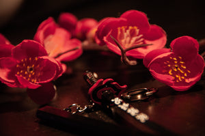 Close-up of a decorative black Demon Slayer Swords! keychain by Incompletegl, featuring intricate silver details and a striking red guard. The keychain is artfully placed among vibrant red flowers with yellow centers on a dark, reflective surface. Bathed in warm lighting, the scene enhances the rich colors of the flowers—making it an ideal accessory for anime enthusiasts.