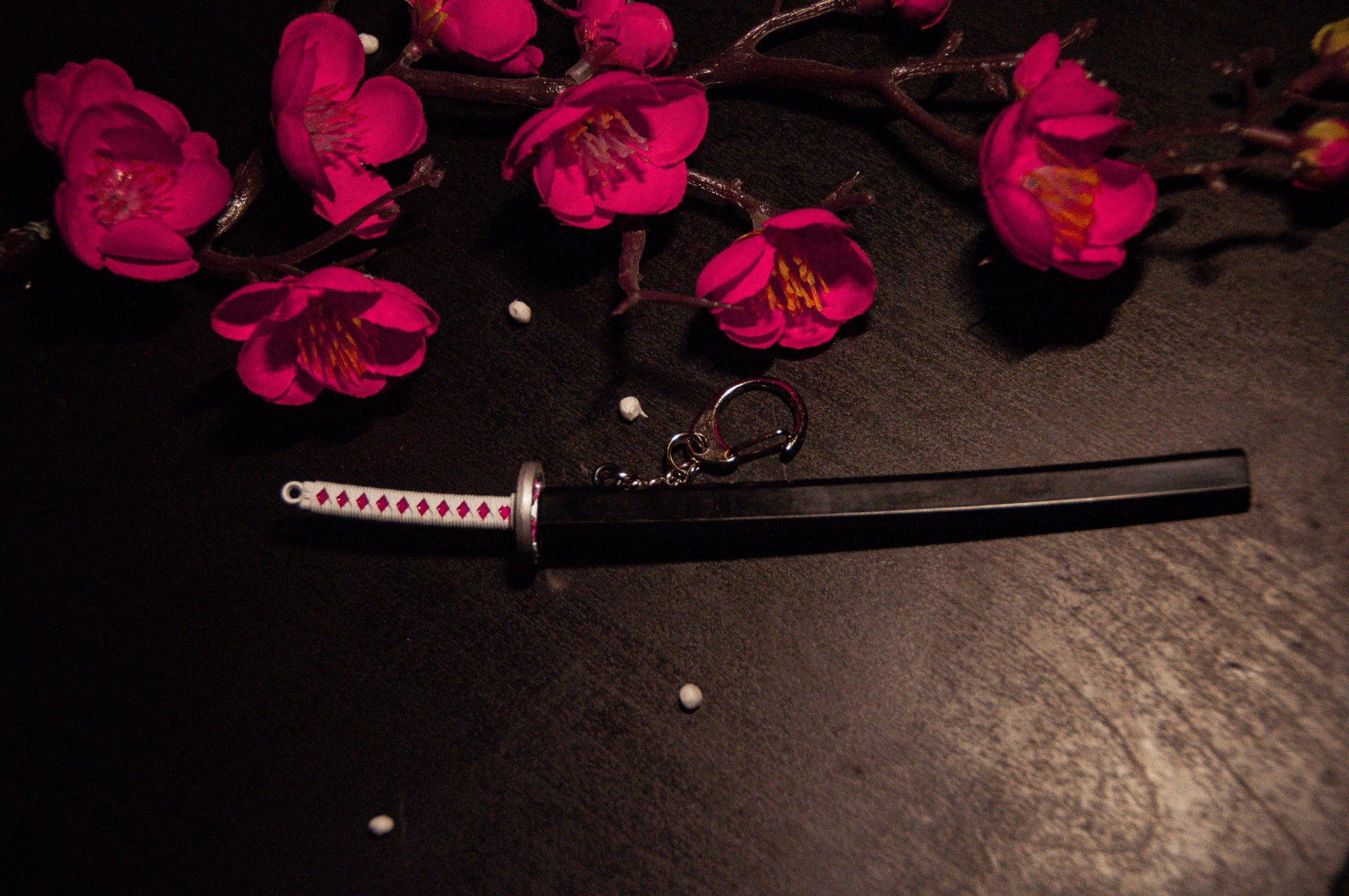 A mini katana keychain, reminiscent of the Demon Slayer Swords! Keychains by Incompletegl, boasts a white handle decorated with pink diamond patterns, lying on a dark surface. To the left, vibrant pink flowers with yellow centers contrast beautifully against the backdrop. Small white beads are scattered around. Perfect for anime enthusiasts.