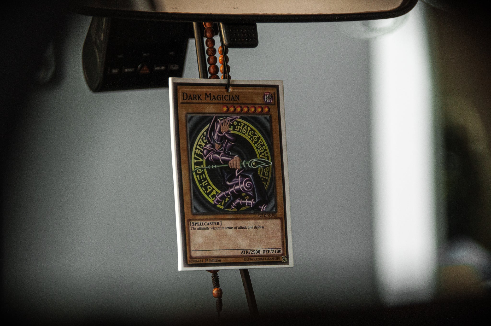 An Incompletegl Dark Magician! air freshener, featuring the spellcaster monster "Dark Magician," is hanging from a rearview mirror inside a vehicle. The air freshener is encased in a protective sleeve, and there is a strand of wooden beads also hanging from the mirror.