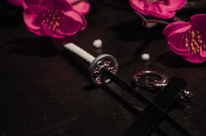 A Demon Slayer Swords! keychain by Incompletegl, featuring a white-handled miniature katana with an intricate flower design on the guard, is elegantly placed on a dark surface. Vibrant pink flowers with yellow centers are scattered around the katana, making it a perfect collectible for anime enthusiasts.