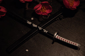 The Demon Slayer Swords! keychains from Incompletegl feature a detailed katana with a white hilt and red diamond accents, resting diagonally on a dark surface. It crosses another black sword adorned with intricate white designs. Surrounding the swords are red flowers, offering a striking contrast of color—an ideal setup for anime enthusiasts who appreciate iconic aesthetics.