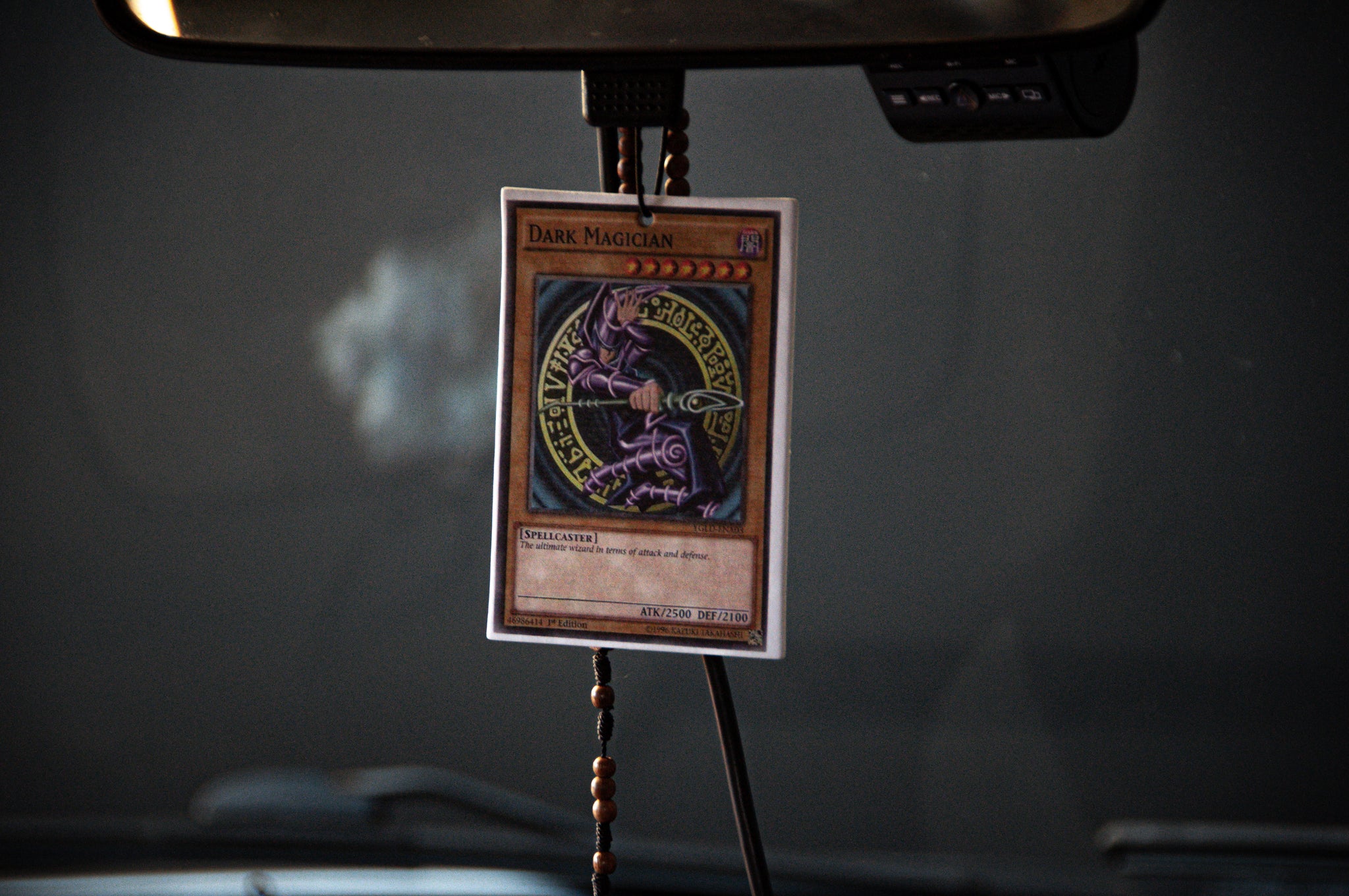 A Dark Magician! | Air Freshener by Incompletegl hangs from a rearview mirror with a beaded string in a car interior. The air freshener features an illustration of the Dark Magician, a powerful spellcaster monster from the beloved trading card game.