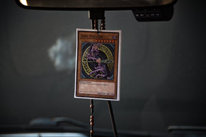 A Dark Magician! | Air Freshener by Incompletegl hangs from a rearview mirror with a beaded string in a car interior. The air freshener features an illustration of the Dark Magician, a powerful spellcaster monster from the beloved trading card game.